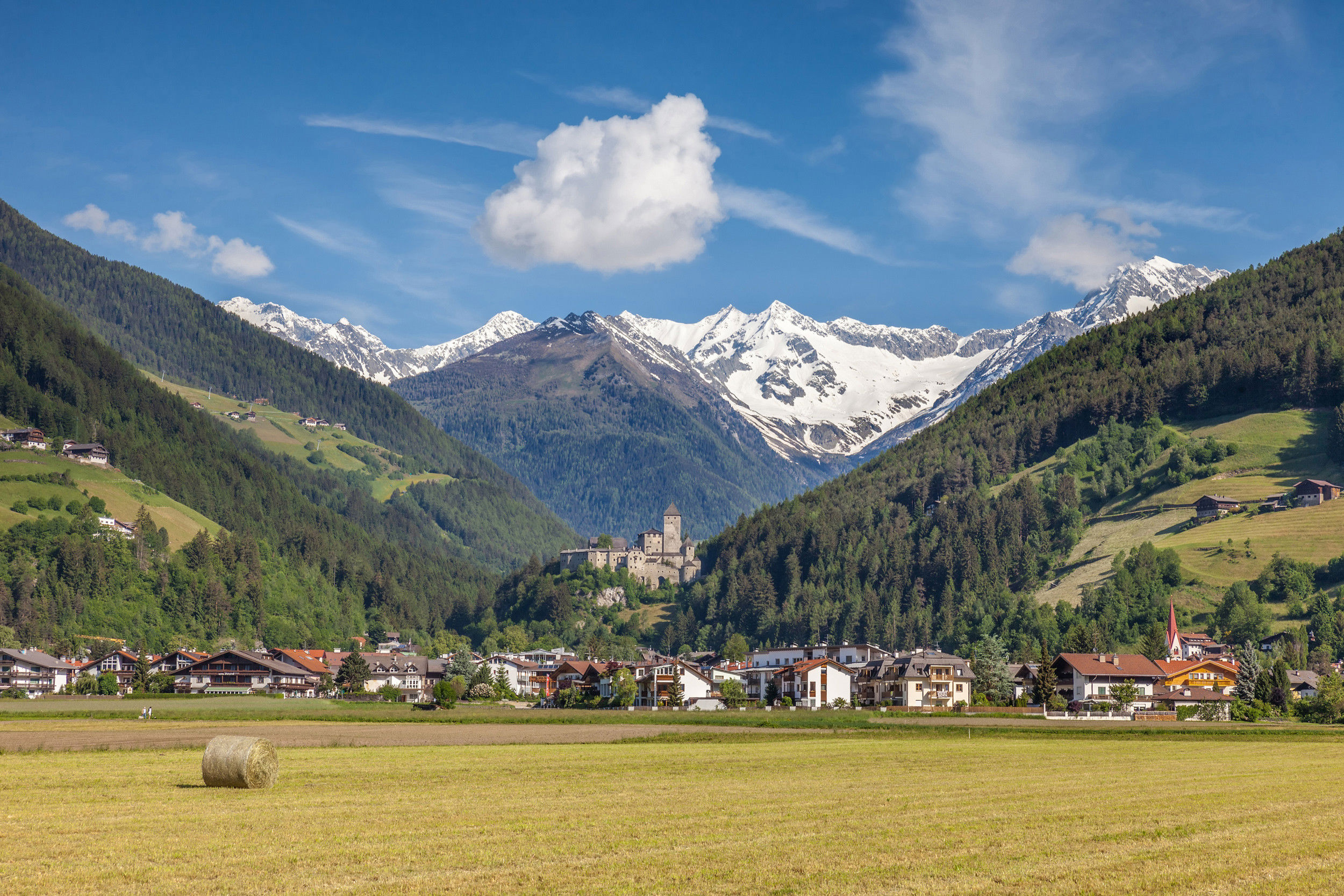View into the valley towards Taufers Castle, framed by fields, houses and snow-covered mountains in the background