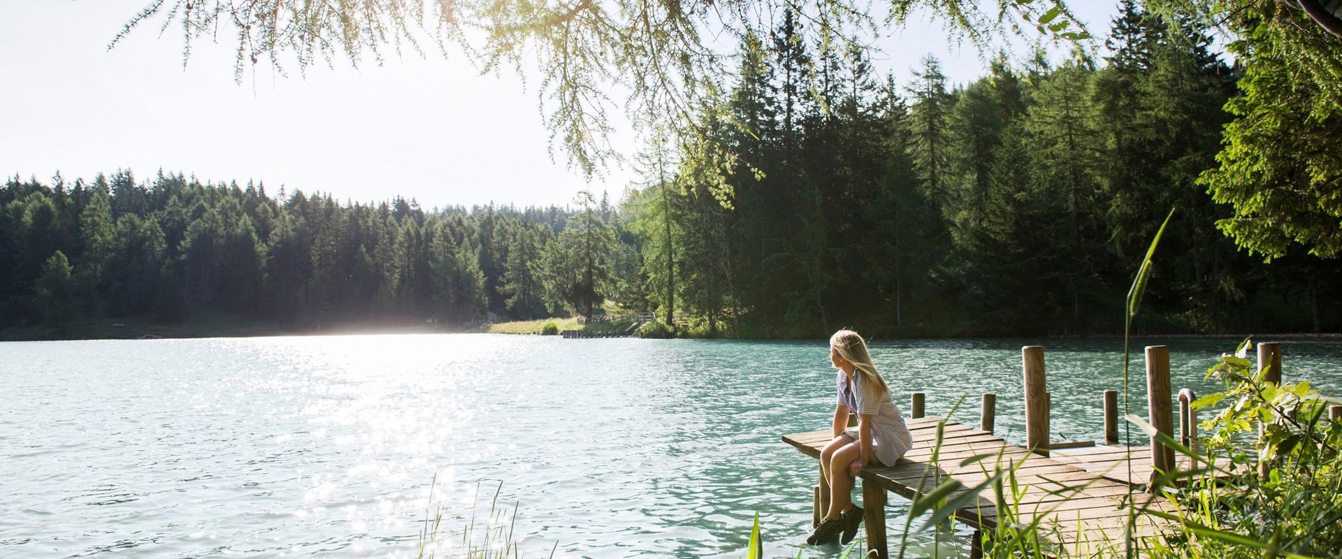 Young girl with blond hair sits on the jetty, on the shore of the lake.