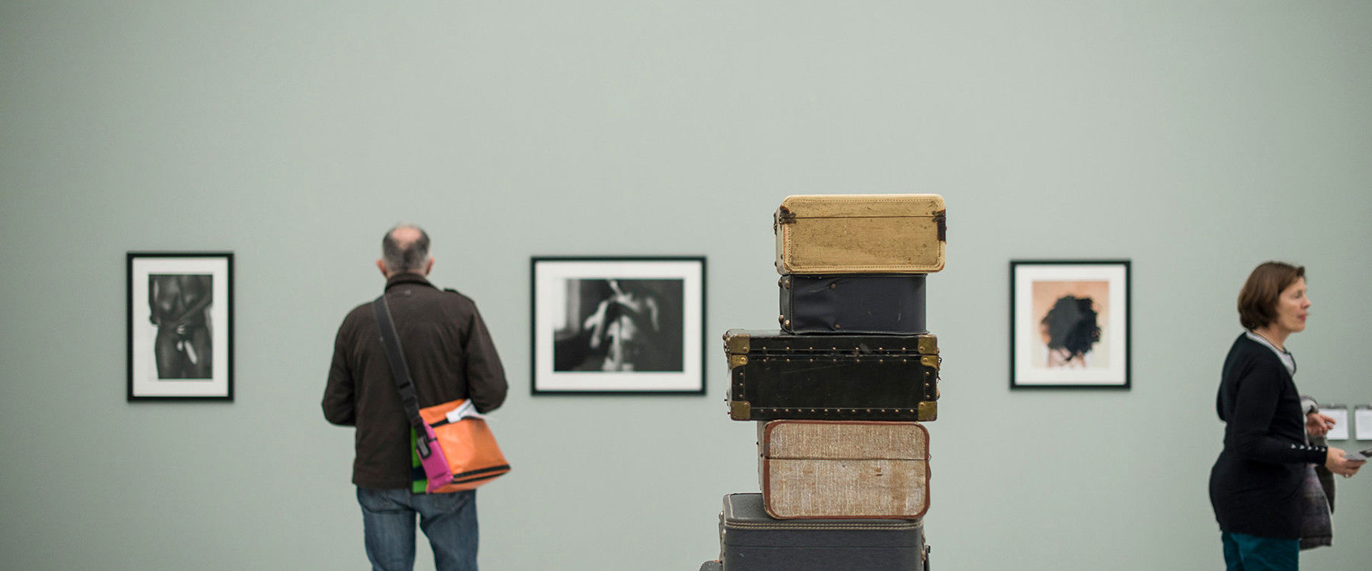 Several old suitcases lie on top of each other in the middle of the room. Pictures hang on the walls and are being examined by 2 visitors.