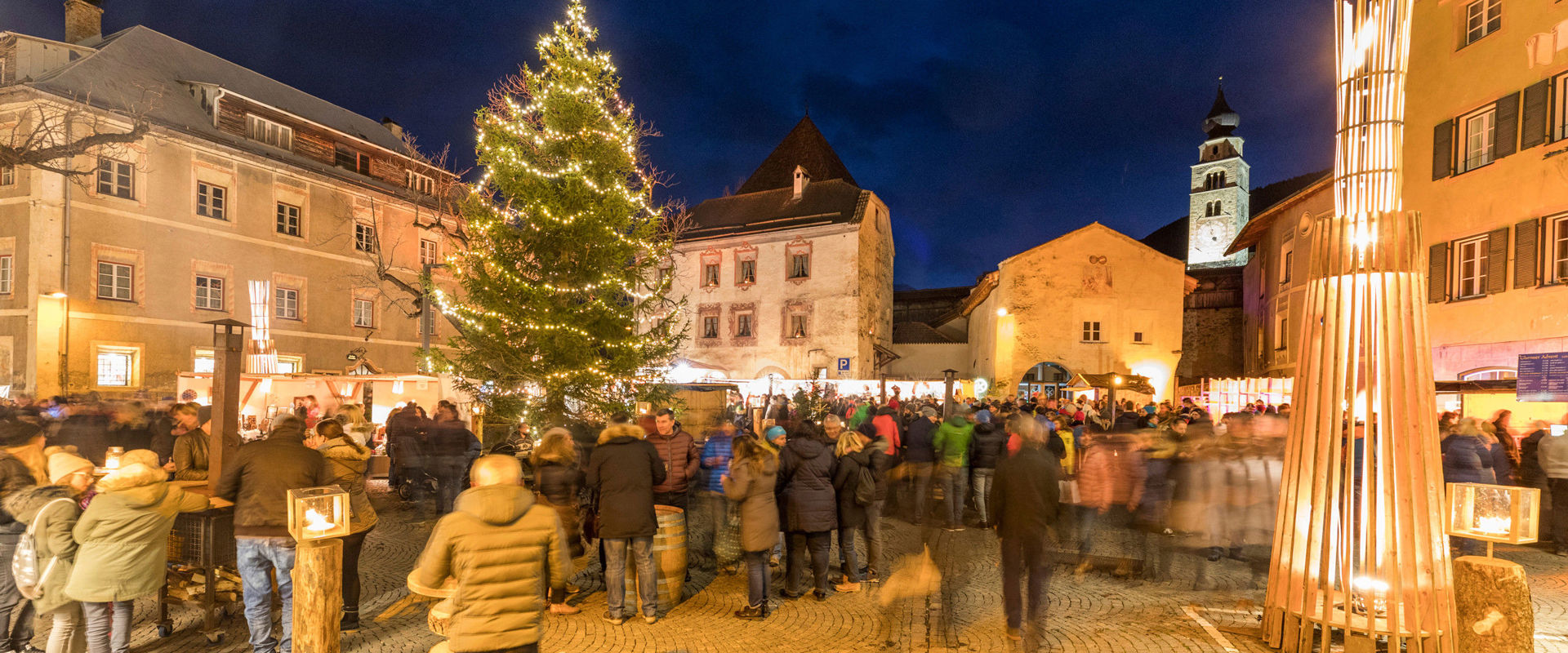 Crowded village square in Glurns with Christmas tree and fairy lights.