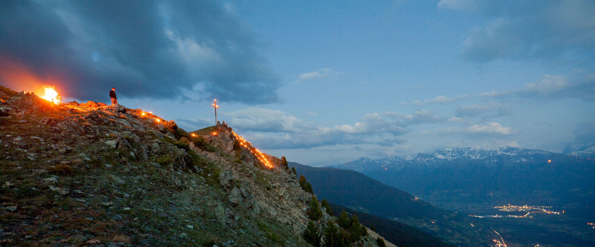 Sacred Heart fire on the mountain top. Man stands next to the summit cross and watches the blazing Sacred Heart fire.