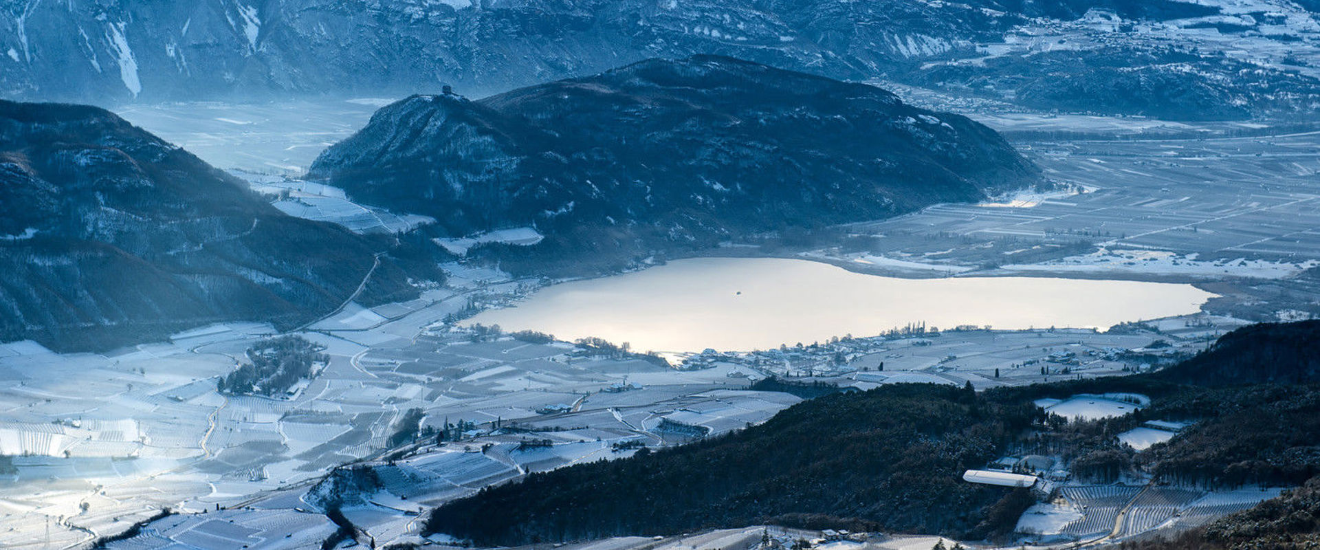 Lake Kaltern in winter Panoramic view of Lake Kaltern in winter. The vineyards, mountains and surrounding nature are covered in snow.
