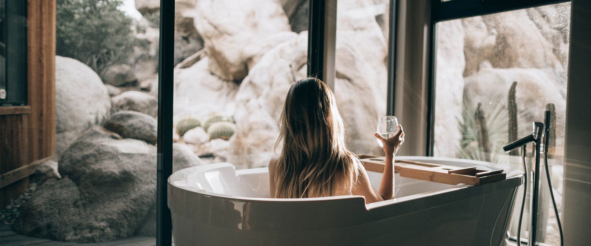 Woman sitting in the bathtub in front of the glazed patio door, drinking a glass of white wine.
