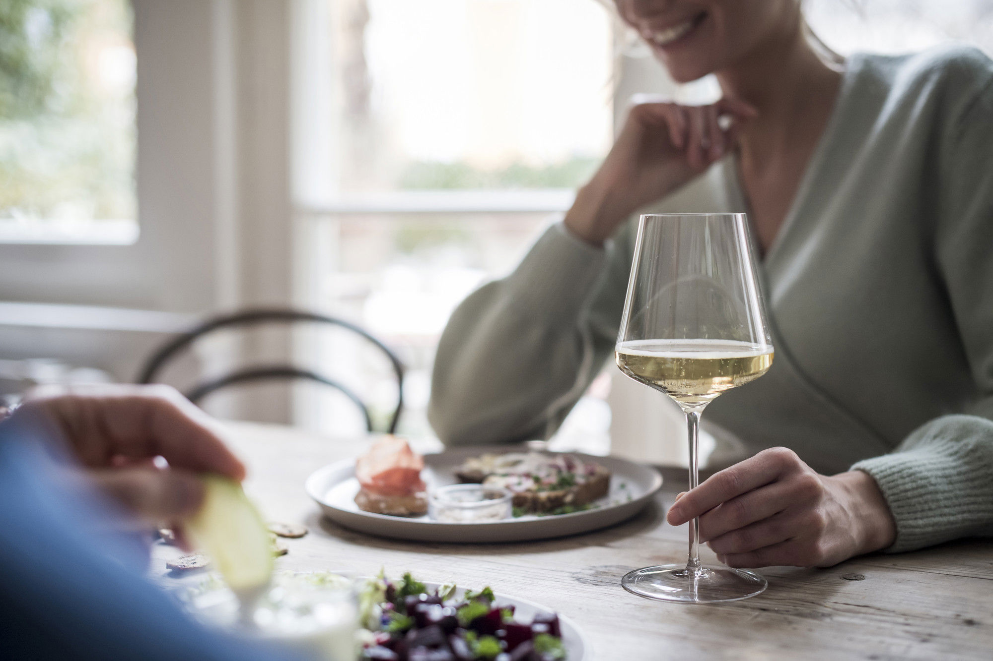 Woman sitting at the table eating and holding a glass of white wine.