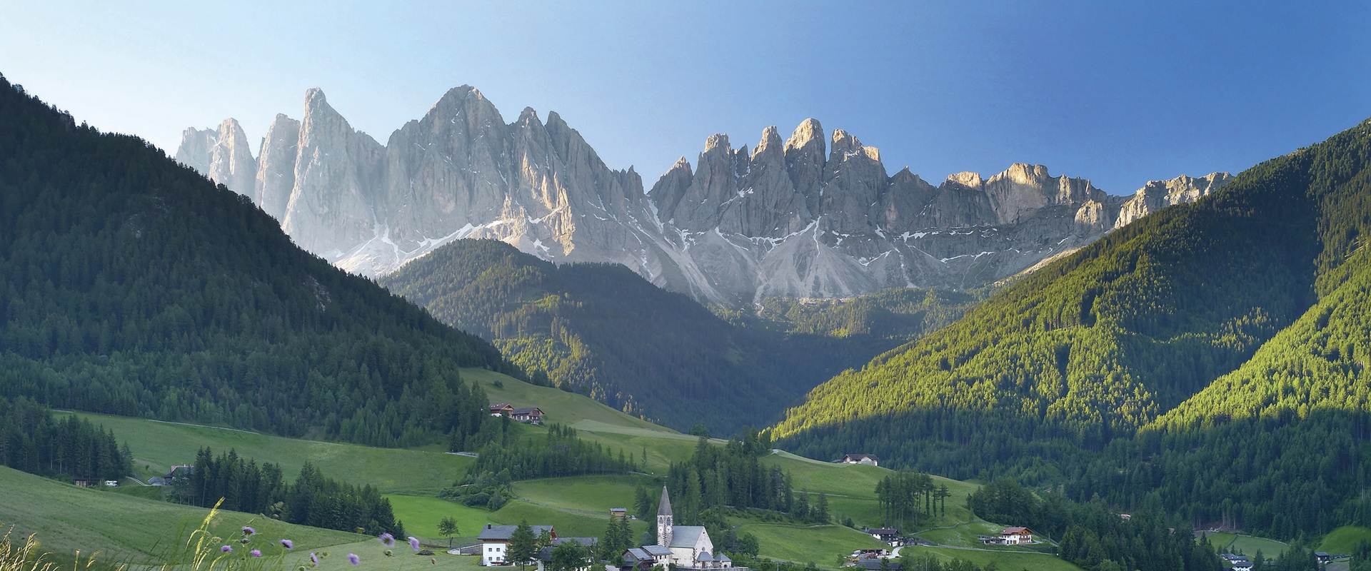Villnöss Valley - Nature Park Puez-Geisler Green mountain meadows, scattered houses and a church in the centre, with grey rocky peaks behind.
